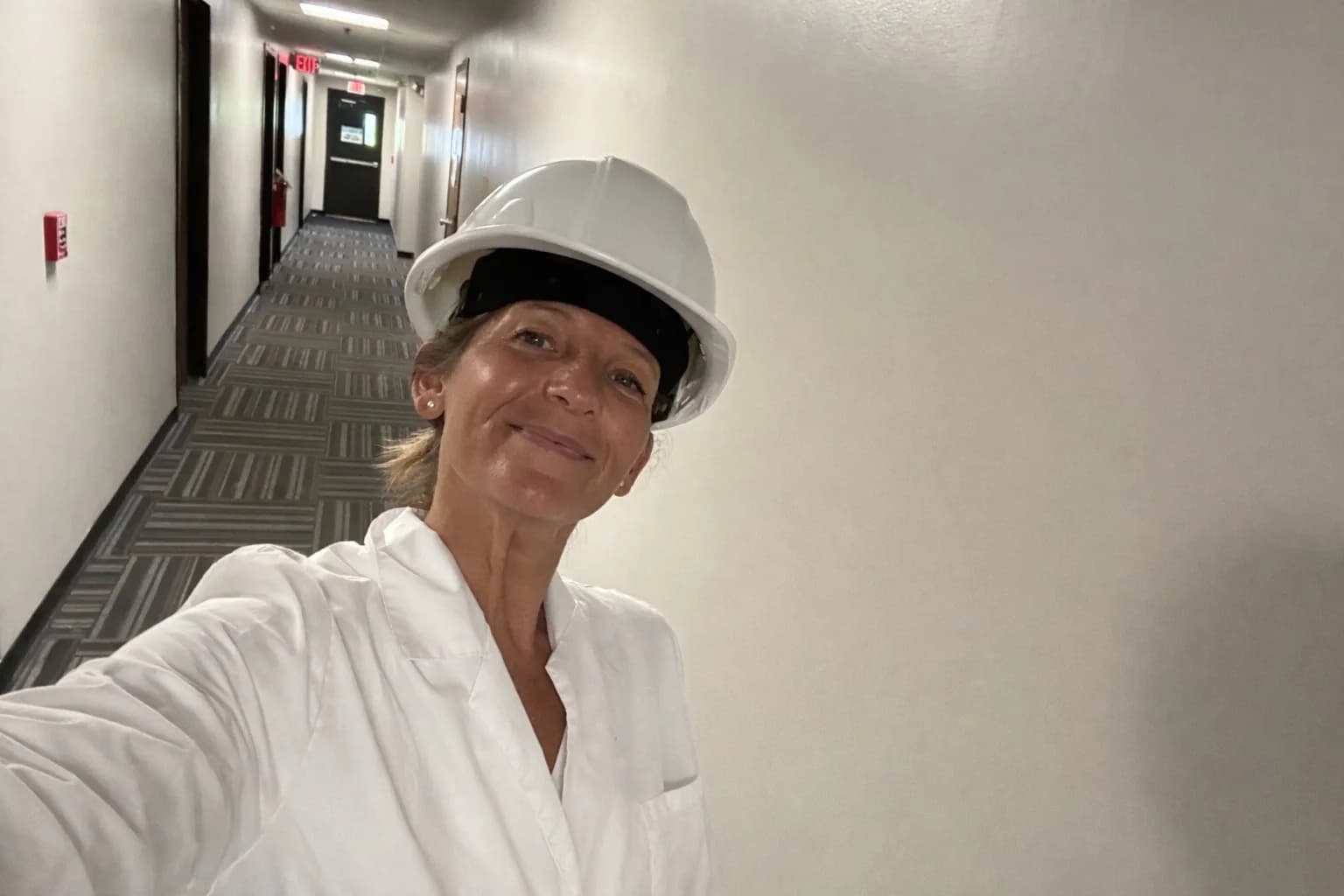 Consultante Lean en casque de chantier blanc et blouse blanche, dans un couloir